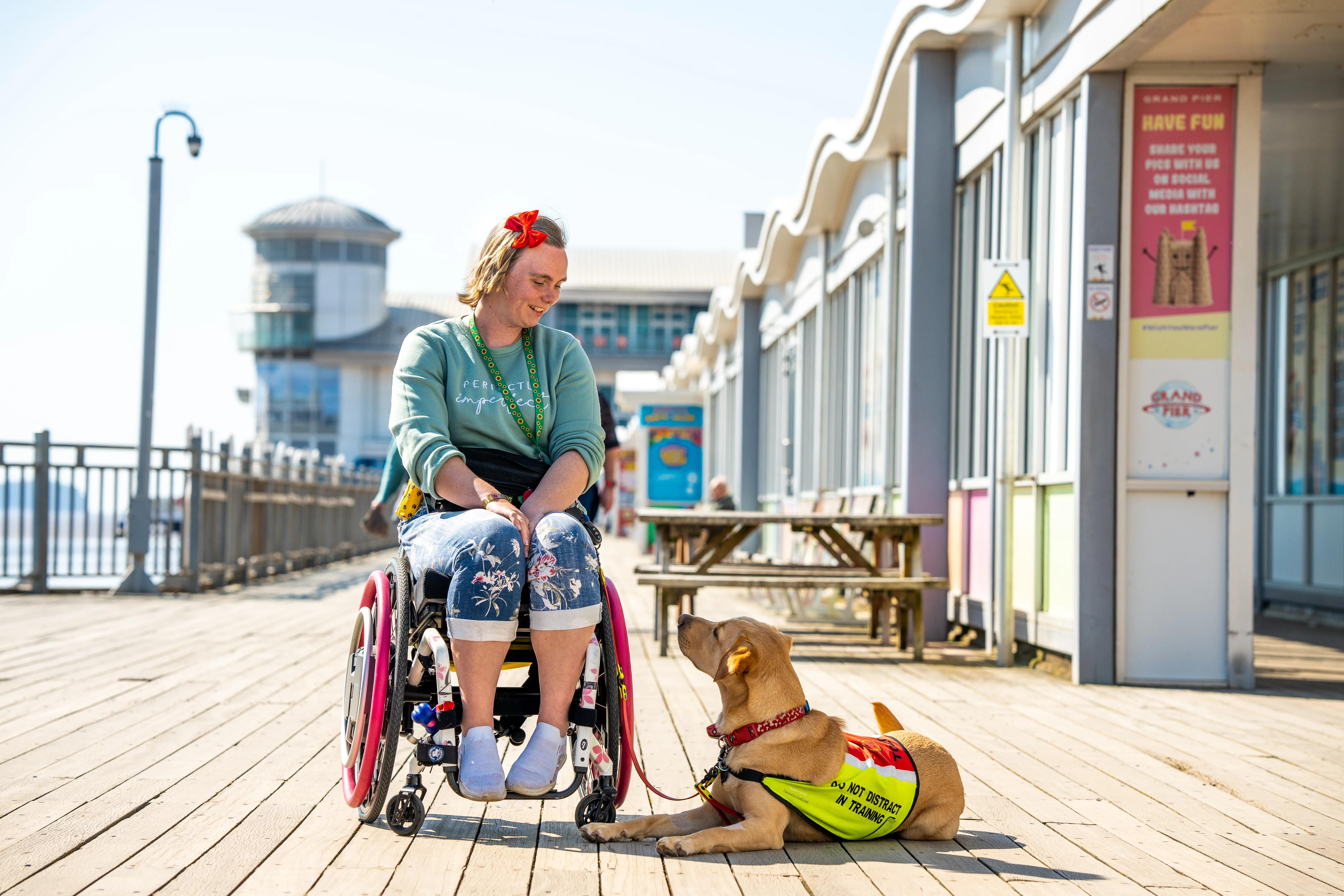 Lady with blond hair and a pale green top and trainers, sat in a wheel chair, looking down at a guide dog. They are on the wooden boardwalk of the Grand Pier.